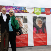 Die drei "Mauthausen-Babys" Eva Clarke, Mark Olsky und Hana Berger Moran stehen lachend vor einer großformatigen Planen-Installation mit ihren Biografien und Fotos am Heldenplatz.