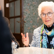 Eva Clarke mit weißem Haar, Brille und buntem Schal sitzt an einem Tisch und gestikuliert beim Sprechen.