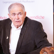 Mark Olsky, mit grauen Haaren im dunklen Sakko, sitzt auf einem Ledersessel und blickt zur Seite. Im Hintergrund ein Roll-up mit dem Logo der Lagergemeinschaft Mauthausen.