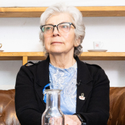 Hana Berger Moran sitzt ruhig auf einem Ledersofa, vor ihr Glaskaraffen. Sie blickt leicht zur Seite.