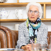 Eva Clarke mit weißem Haar, Brille und farbigem Schal, sitzt auf einem Ledersofa, vor ihr mehrere Gläser. Sie schaut konzentriert nach vorne.