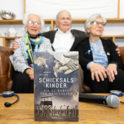 Das Buch "Schicksalskinder – Die KZ-Babys von Mauthausen" steht im Fokus, im Hintergrund sitzen die "Mauthausen-Babys" auf einem Sofa.