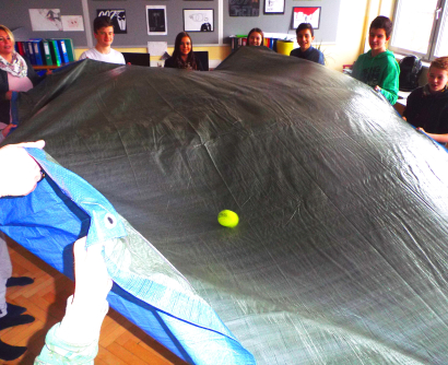 Gruppenaufnahme eines MKÖ-Workshops. Jugendliche und Trainerin führen eine Übung mit Fallschirmtuch und Tennisball durch.