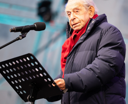 Paul Lendvai, in Winterjacke und rotem Schal, spricht beim Fest der Freude 2025, das Mikrofon und Notenpult vor sich.